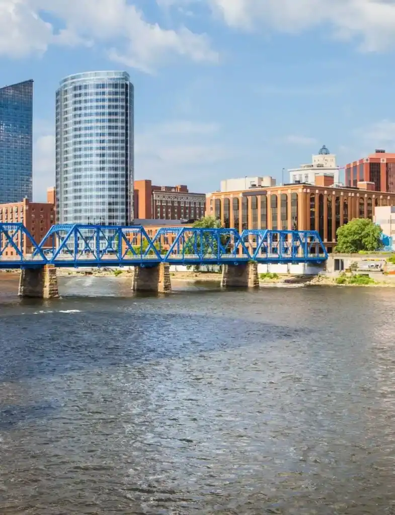 Blue truss bridge spanning river with modern and historic buildings—Grand Rapids cityscape featured as a key Grey Cap service location.