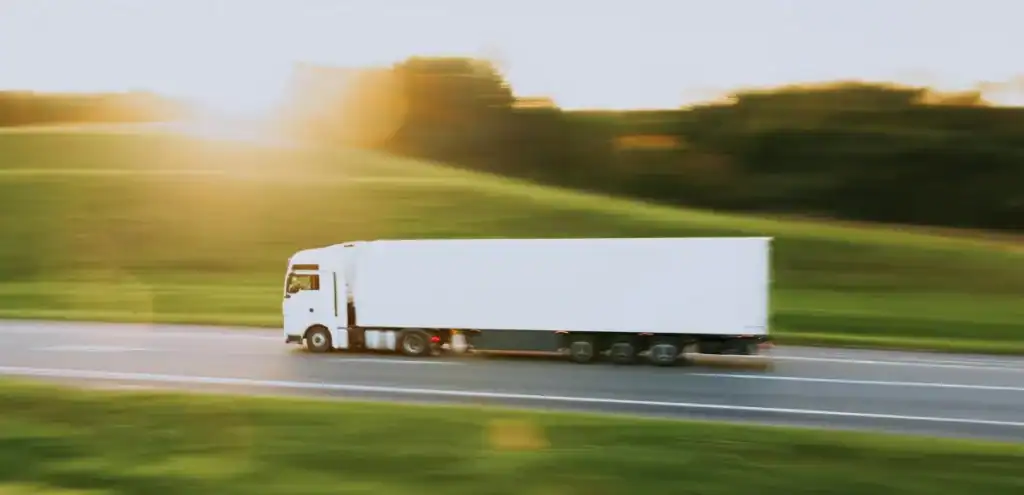 White semi-truck driving at high speed on a highway through grassy hills during sunrise or sunset—illustrating Last Mile transport service at Grey Cap.