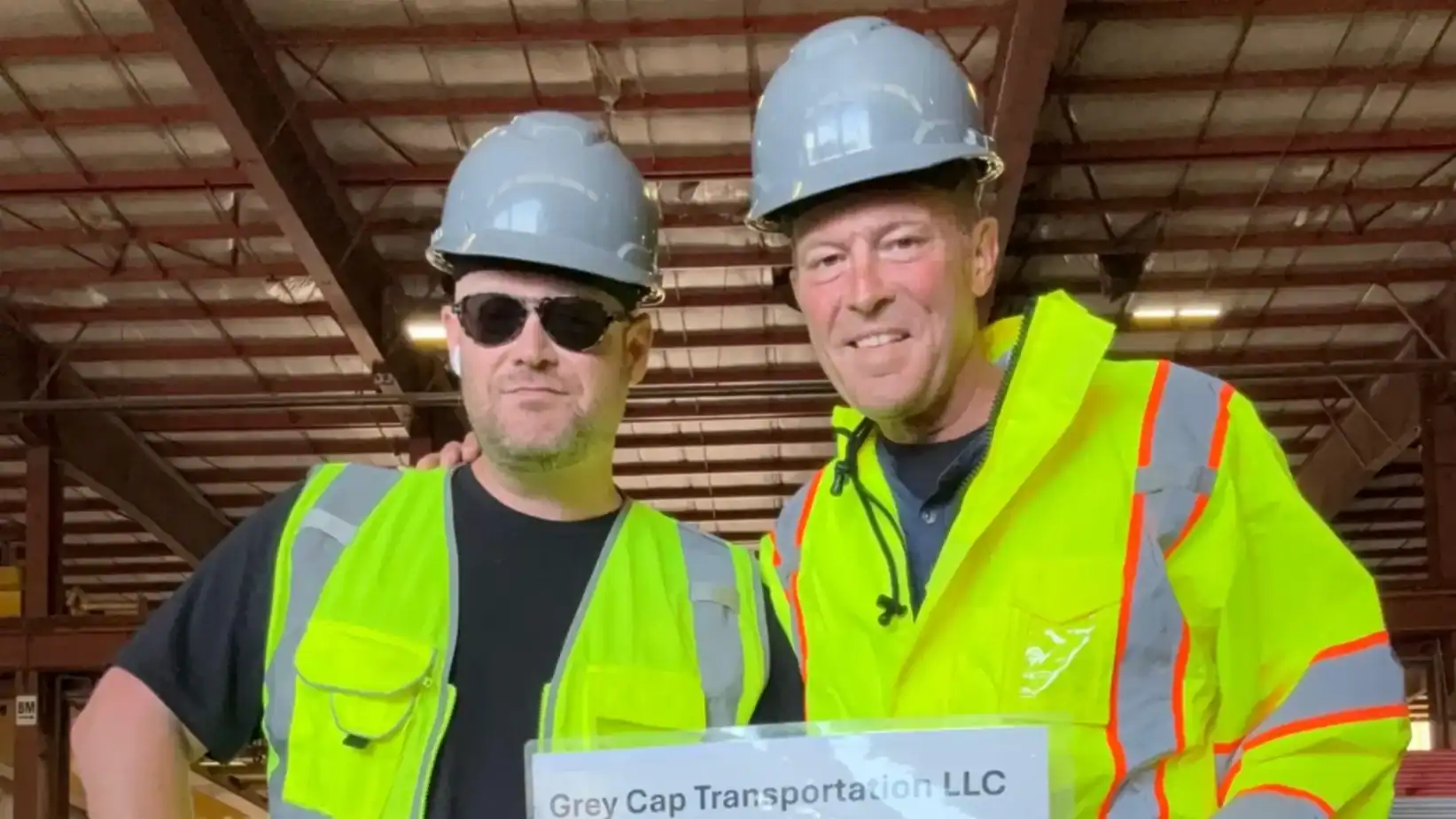 Two workers in hard hats and hi-vis vests in warehouse holding Grey Cap Transportation LLC sign—safety-focused team.